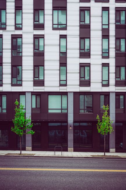 A modern multi-storey apartment building with light-colored brick facade and large glass balcony doors, situated in a landscaped area with paved walkways, small trees, and patches of grass. The balcon